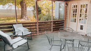 A covered outdoor seating area with a table and chairs, a wooden fence, and a view of trees and a building in the background. - Nashville Haven Hideaway- Cozy 3 Bedroom House (Nashville)