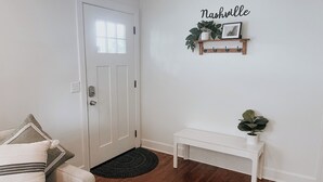 A clean, white-walled room with a door, a small white bench, a black rug, and a wall-mounted shelf with plants and a picture. - Nashville Haven Hideaway- Cozy 3 Bedroom House (Nashville)