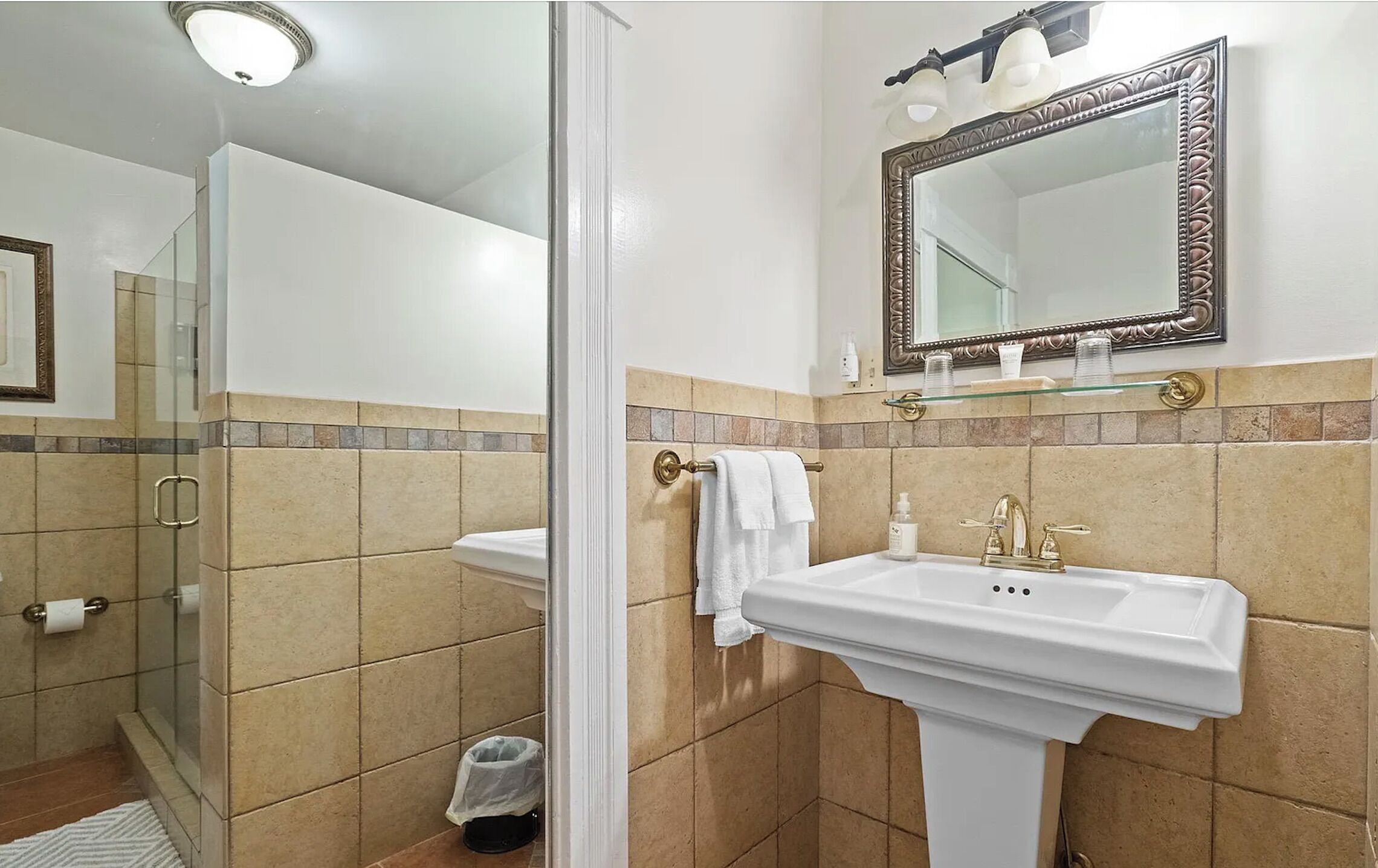 A bathroom with a glass shower enclosure, a pedestal sink with a gold faucet, and a large mirror with a decorative frame.