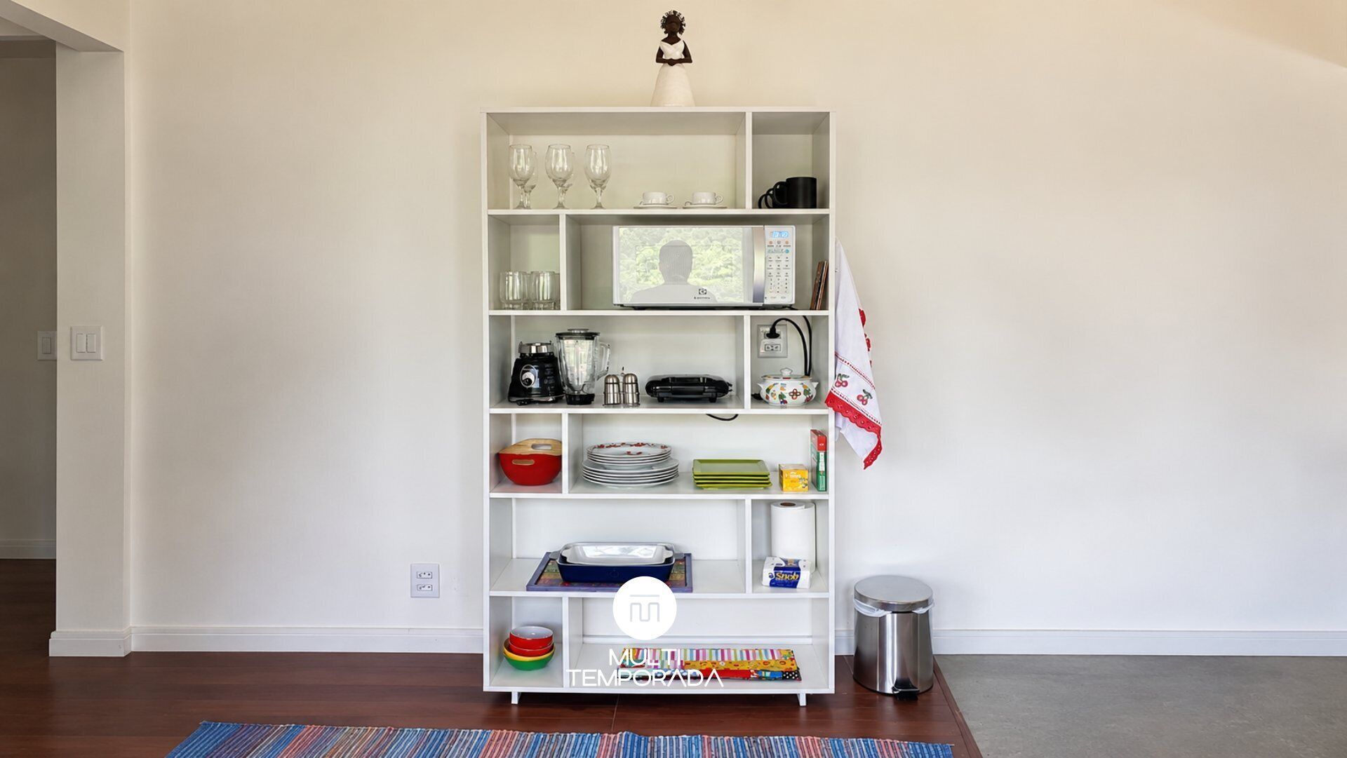 A white shelving unit with kitchenware, a microwave, and a trash can.