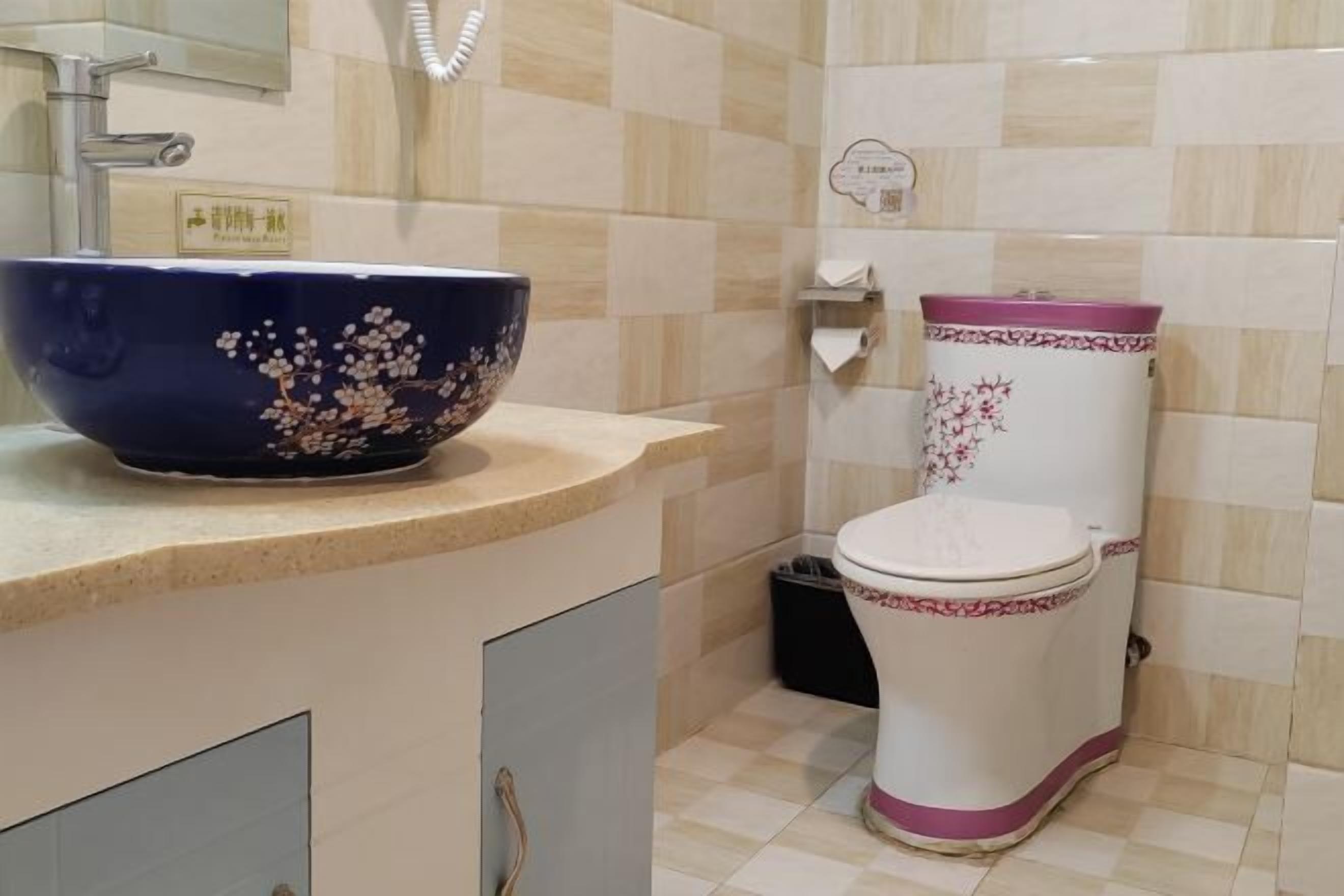 A bathroom with a blue ceramic basin, a white toilet with floral trim, and beige tiled walls.