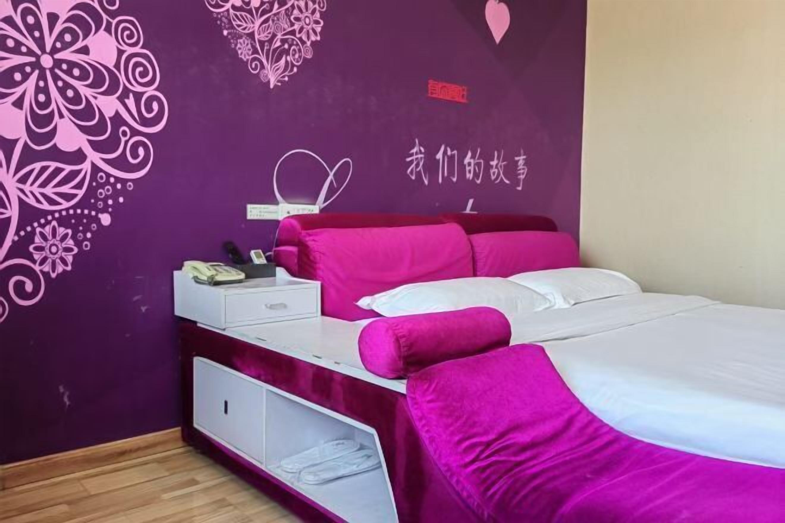 A hotel room with a purple wall, a bed with pink bedding, a white nightstand, and a decorative wall design.
