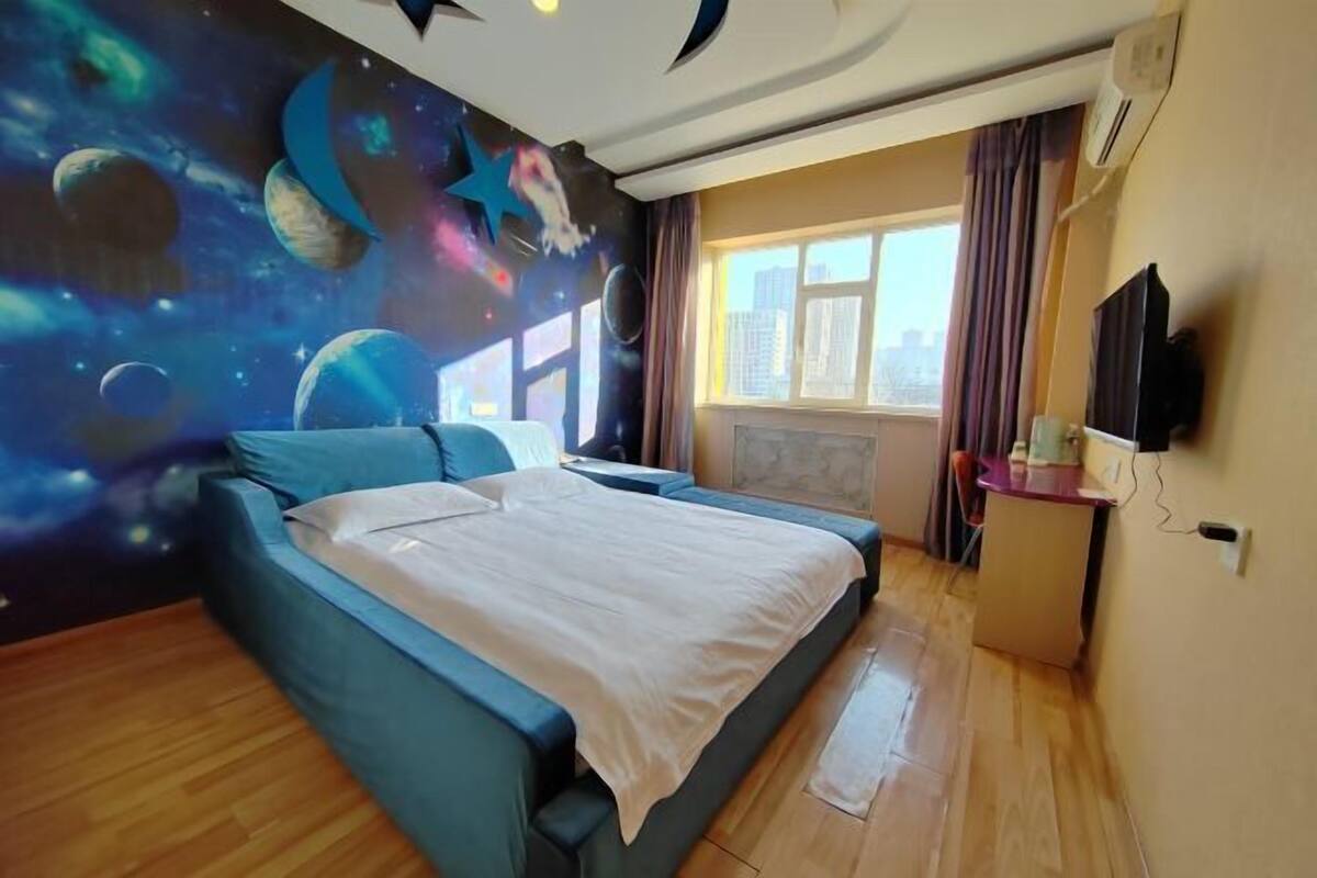 A bedroom with a bed, a wall mural of space, a window with curtains, and a TV on the wall.