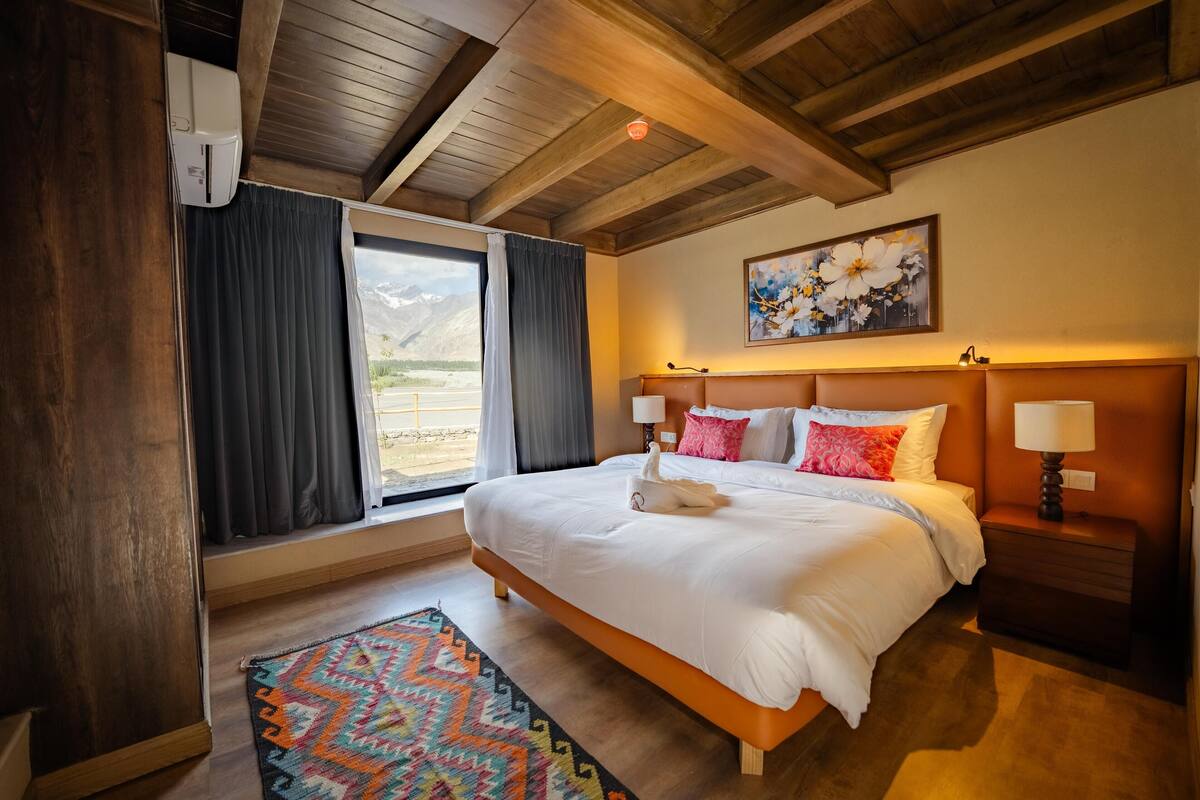 A hotel room with a large bed, a wooden ceiling, a painting on the wall, and a window with curtains.