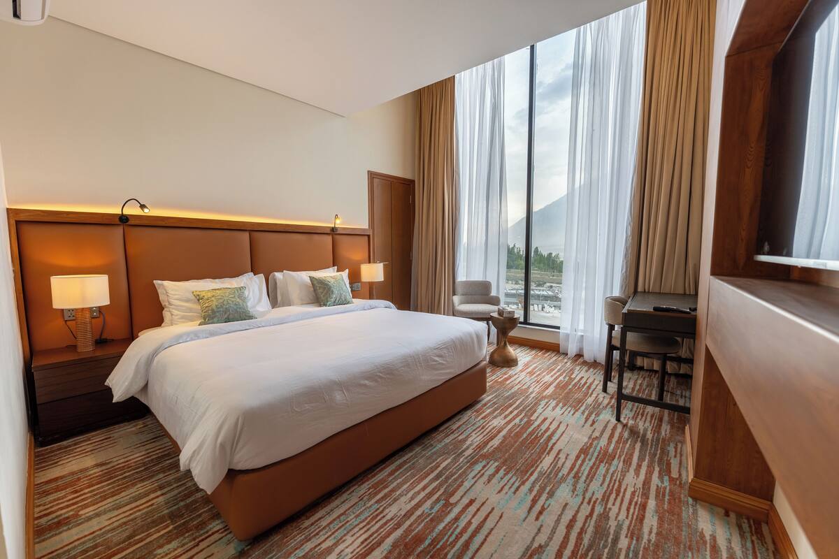 A hotel room with a large bed, two bedside lamps, a desk with a chair, and a large window with curtains.