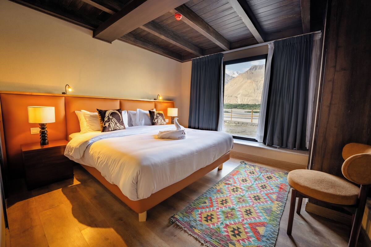 A bedroom with a large bed, wooden headboard, bedside lamps, a colorful rug, a chair, and a view of mountains through the window.