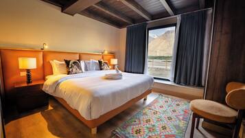 A bedroom with a large bed, wooden headboard, bedside lamps, a colorful rug, a chair, and a view of mountains through the window.