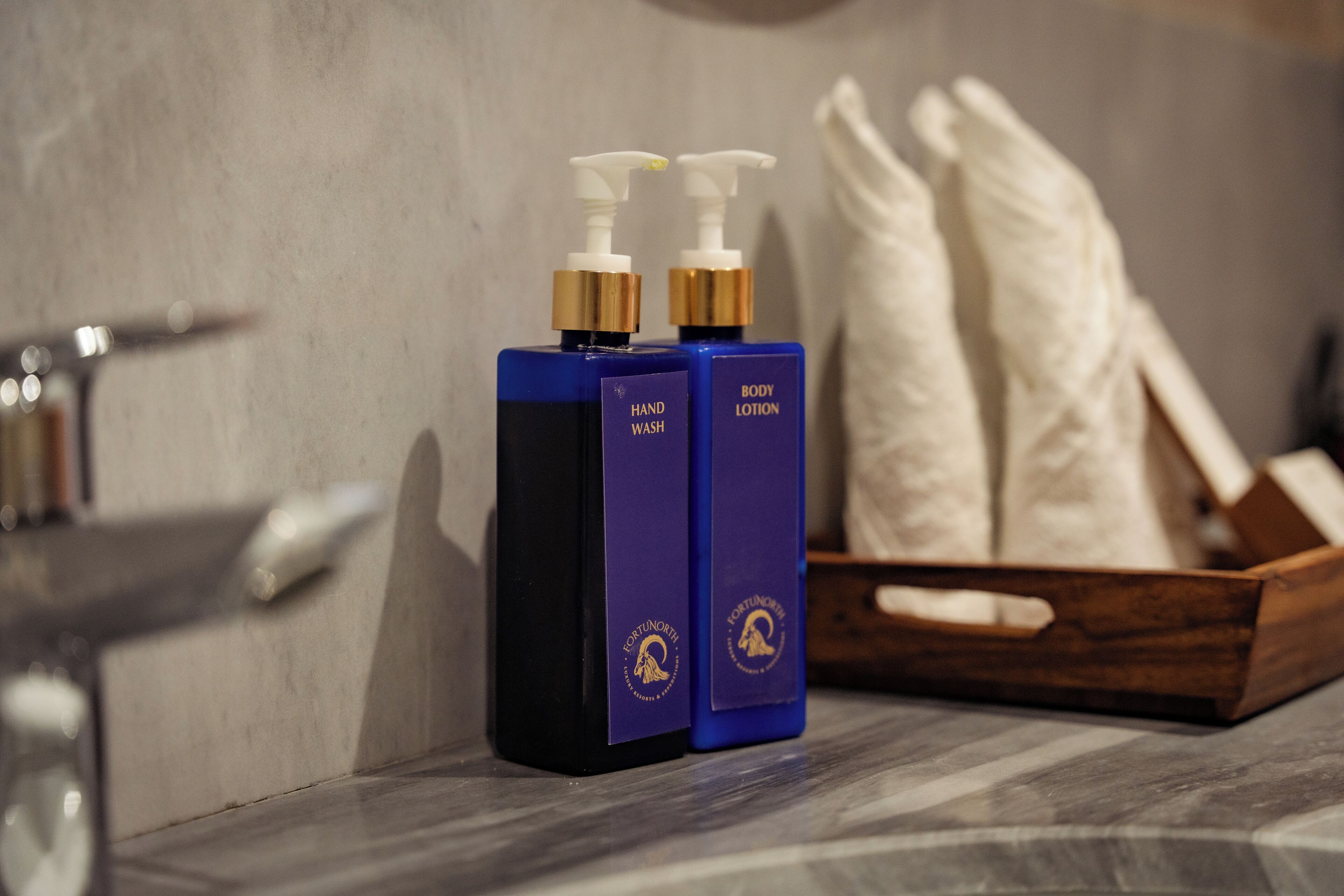 Two bottles of hand wash and body lotion with pumps, placed on a marble surface next to a wooden box with towels.