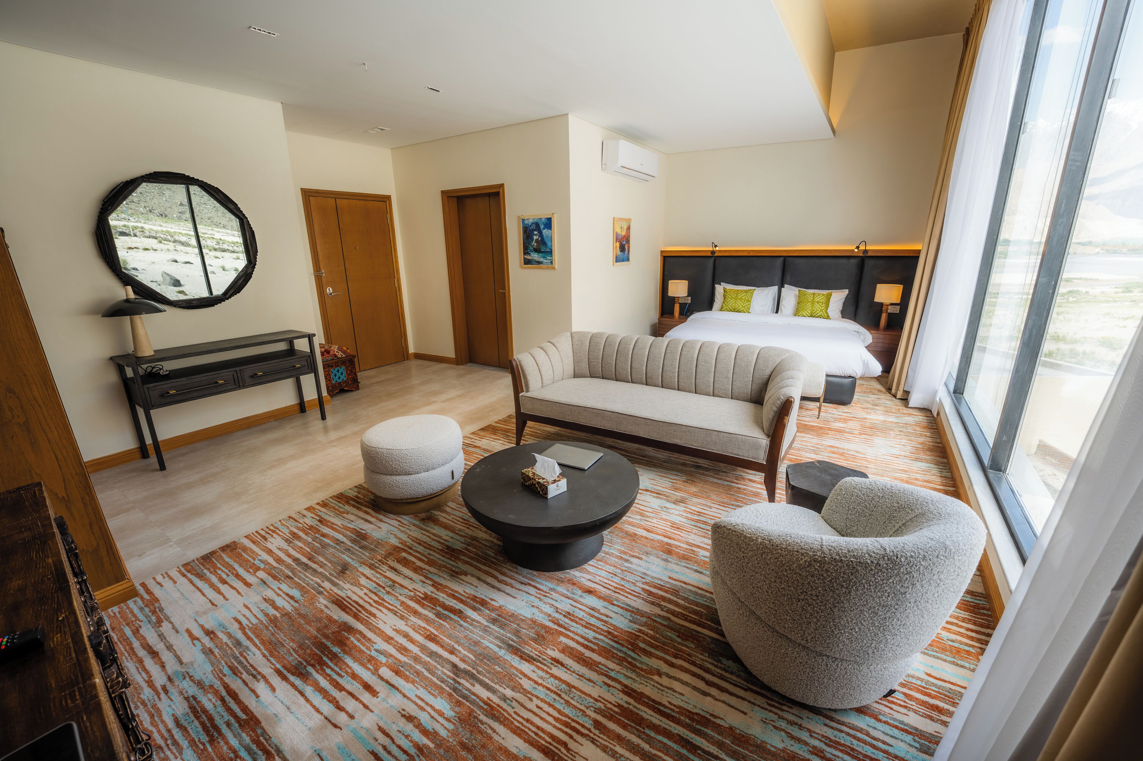 A modern hotel room with a large bed, a sofa, a round coffee table, and a seating area with a chair. There is a mirror on the wall, a small table with a lamp, and a view of the outdoors through the window.