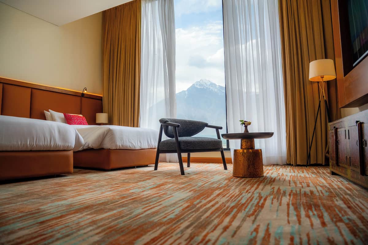 A hotel room with a bed, a chair, a small table, and a view of a mountain.