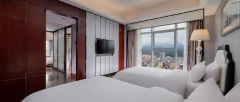 A hotel room with two beds, a TV, a large window with a city view, and a door leading to another room.