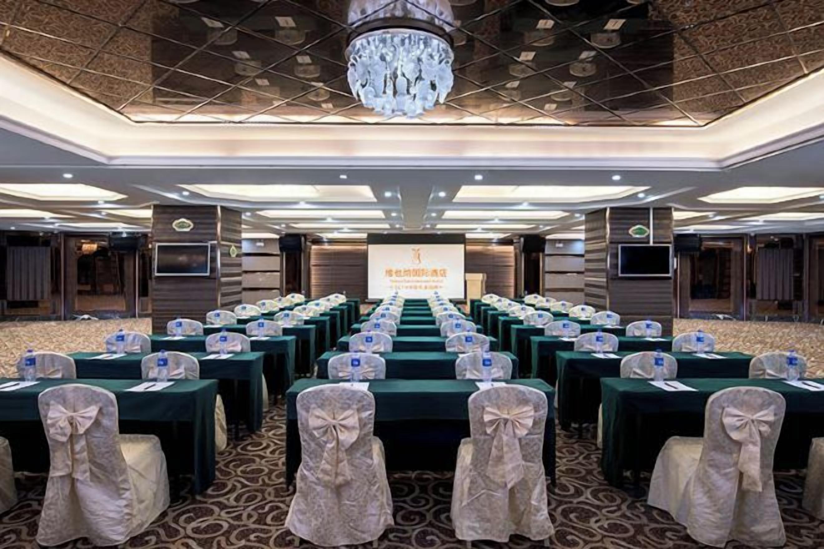 Une salle de conférence avec des rangées de tables et de chaises, un grand écran à l’avant et un lustre.