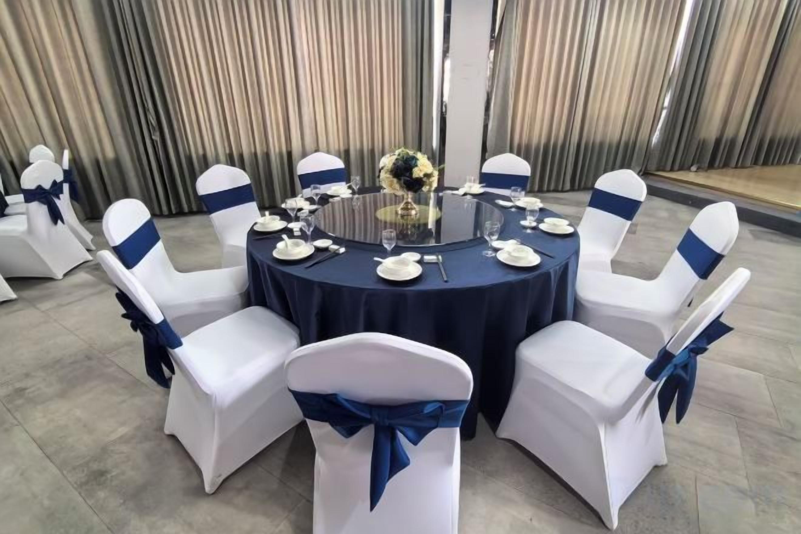 Une table ronde recouverte d’une nappe sombre, entourée de chaises blanches ornées de rubans bleus, préparée pour un événement officiel.