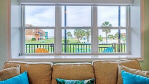A room with a large window overlooking a courtyard with palm trees and a pool. There is a sofa with cushions in the foreground. - Sleeps 22 3 Condos in Beachfront Complex w/ Pool, Pickleball Island Retreat (Port Aransas)