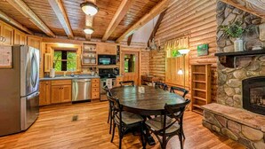 A rustic cabin interior with a wooden dining table, chairs, a stone fireplace, and a kitchen area with appliances. - Lakefront Log Cabin at Lake Anna, Dock, Deck (Bumpass)