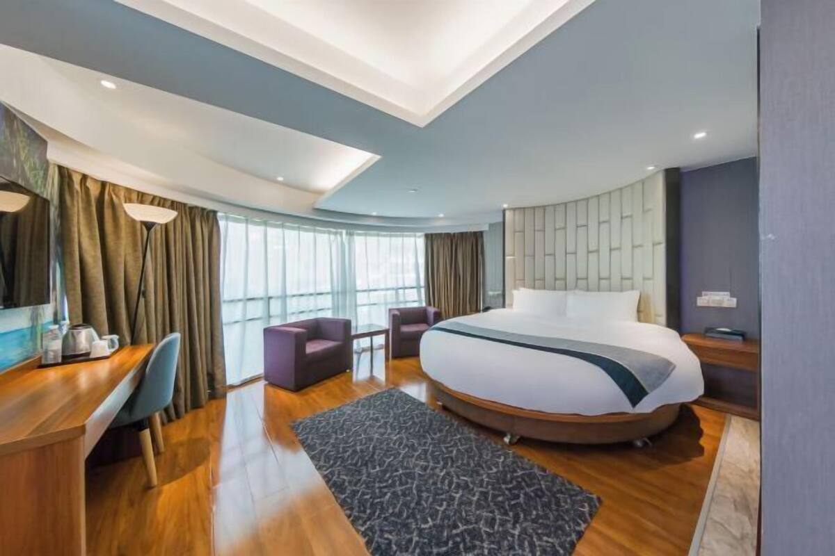 A modern hotel room with a large bed, a desk with a chair, a small seating area, and a large window.