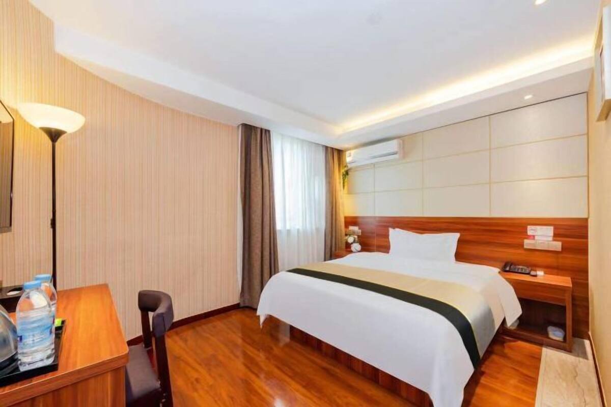 A hotel room with a wooden bed, a desk with a lamp, a chair, a bedside table with a phone, and a wall-mounted air conditioning unit.