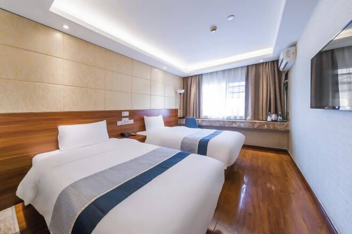 A hotel room with two beds, a wooden headboard, a large window with curtains, a flat-screen TV, and a minibar.