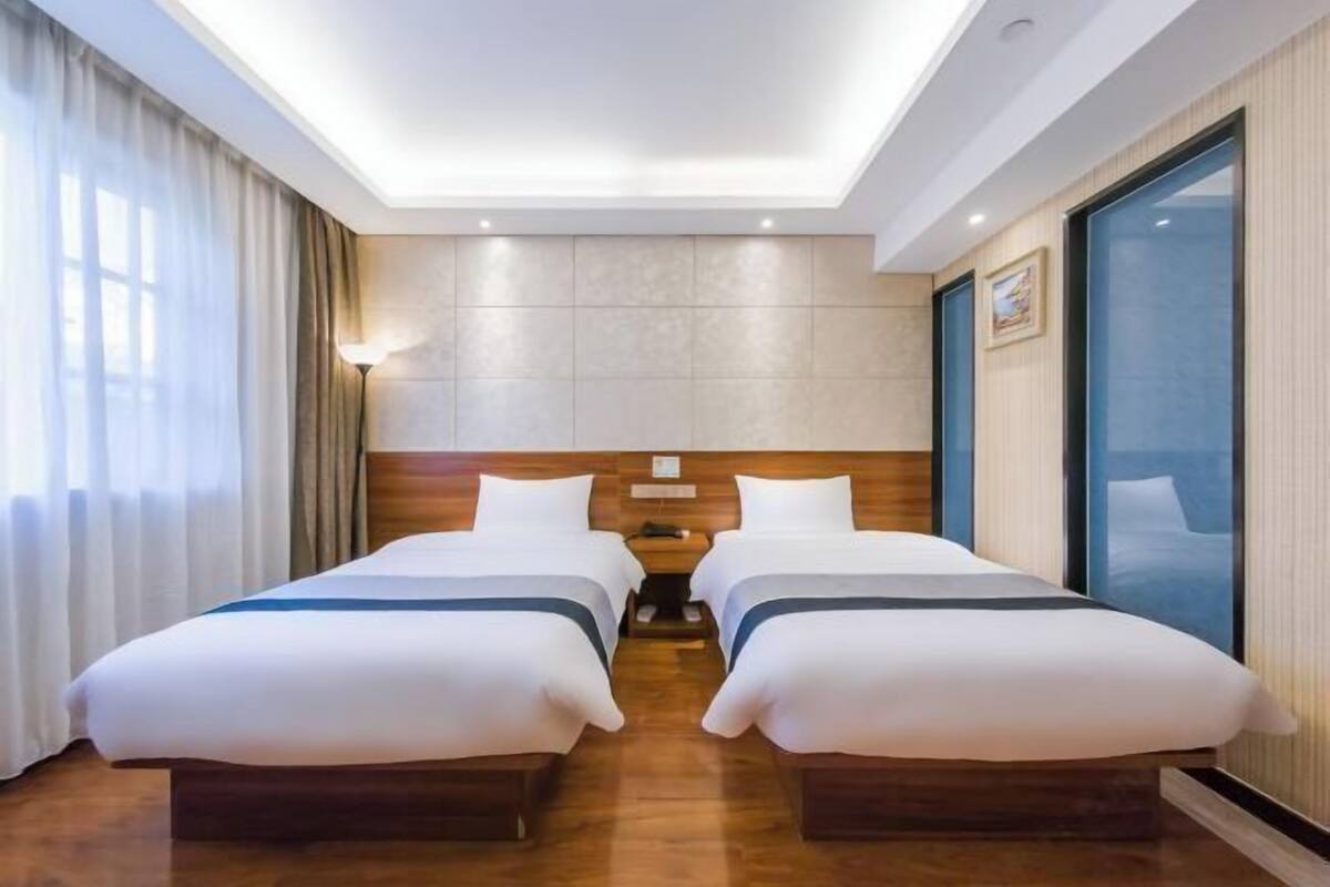 A hotel room with two beds, a nightstand, a lamp, a large window, and a ceiling light.
