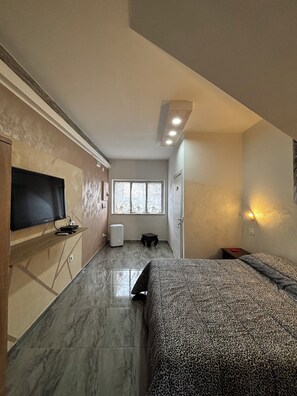 Une chambre d’hôtel de taille réduite, comprenant un lit, un téléviseur fixé au mur, une petite table et une fenêtre laissant entrer la lumière naturelle.
