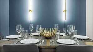 Mesa de comedor preparada para seis personas, con un cuenco decorativo en el centro, dos lámparas colgantes y una pared de color azul oscuro al fondo.