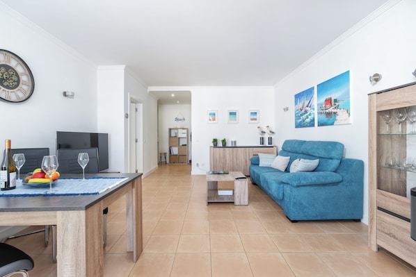 A modern living room with a blue sofa, a dining area with a wooden table, and a kitchen in the background.