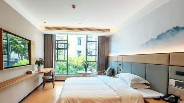 A modern hotel room with a large bed, a desk with a chair, and a large window offering a view of greenery outside. - Taihe Jinghu Hotel (Taihe)