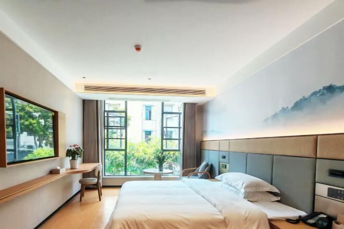 Habitació d'hotel moderna amb un llit gran, una taula de treball amb una cadira i un finestral ampli que ofereix vistes a la verdura.