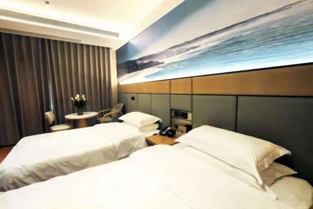 A hotel room with two beds, a desk, a chair, a small table with a plant, and a painting of a seascape on the wall.