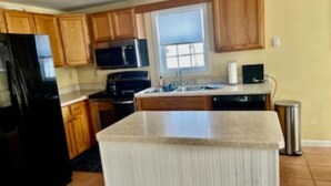 A kitchen with wooden cabinets, a black refrigerator, a microwave, a stove, and a sink. - Charming  3B/R  Cottage on Malletts Bay (Colchester)