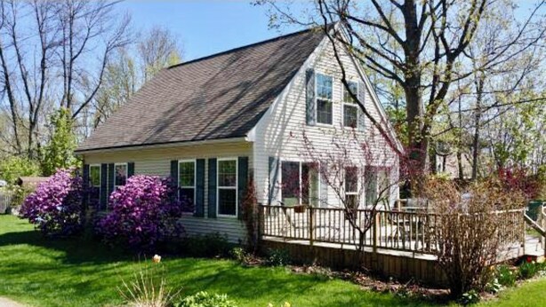 A two-story house with a gabled roof, a front porch, and a small garden with purple flowers. - Charming  3B/R  Cottage on Malletts Bay (Colchester)