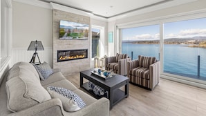A modern living room with a fireplace, comfortable seating, and a large window offering a scenic view of a lake. - Coastal View Sunset at SookePoint Cottages (Sooke)