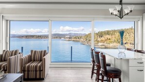 A modern kitchen with a large window overlooking a lake, a dining area with striped chairs, and a bar stool. - Coastal View Sunset at SookePoint Cottages (Sooke)