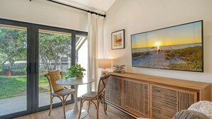 Una sala moderna con un ventanal grande, un comedor con mesa y sillas, una consola de madera y un cuadro grande de un atardecer sobre el océano.