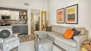 Una sala moderna con un sofá seccional gris, un reposapiés gris y un sillón estampado. Se observa una cocina al fondo con gabinetes grises y un microondas.