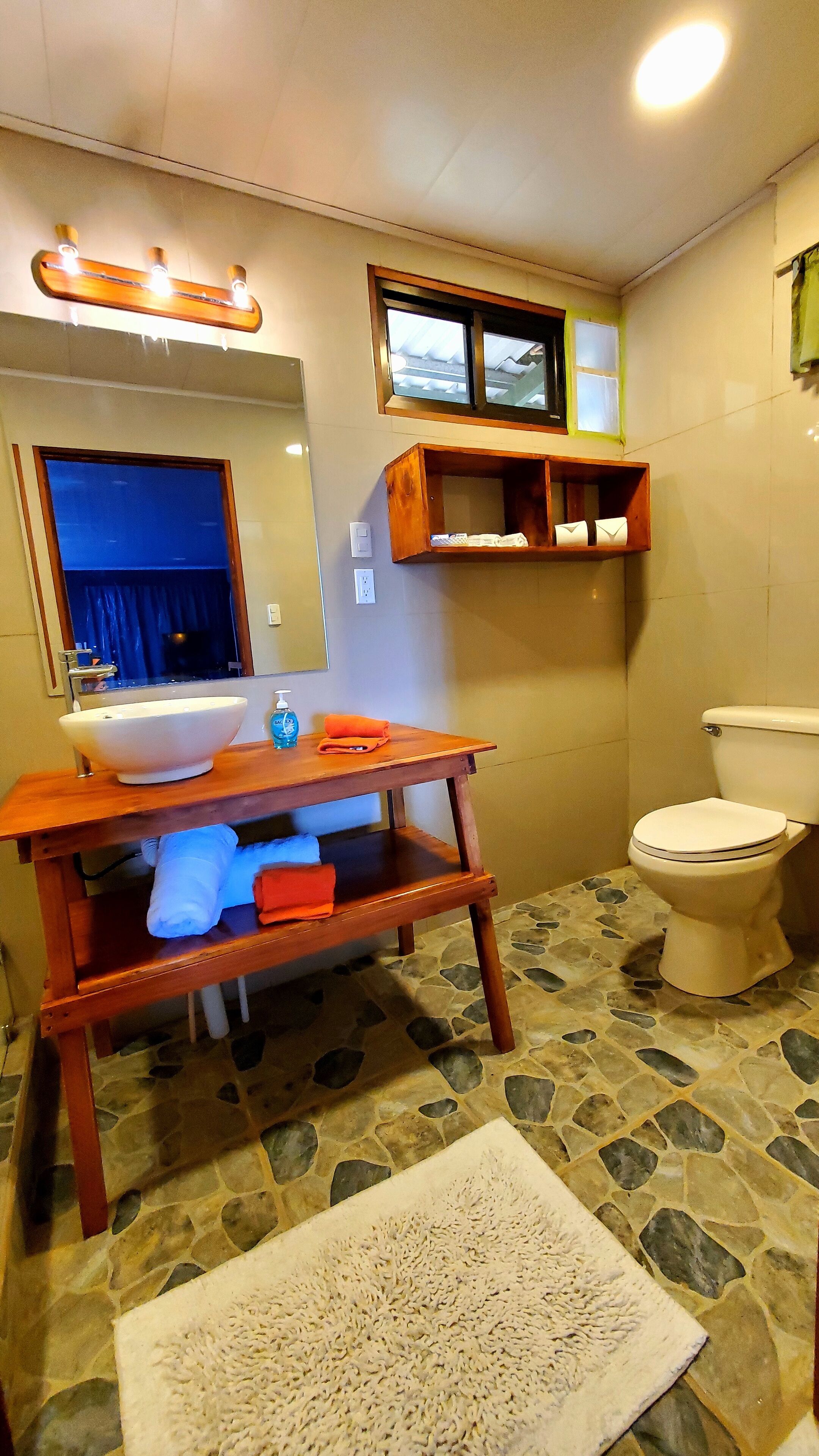 Une salle de bain avec un meuble-lavabo en bois, un grand miroir, des toilettes et un sol carrelé en pierre.