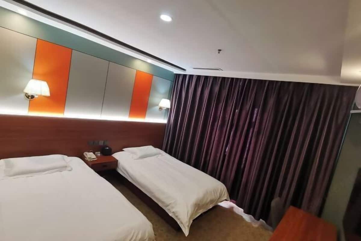 A hotel room with two single beds, a wooden headboard, a nightstand with a lamp, and a large window with curtains.