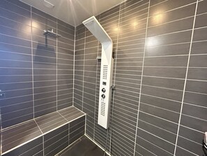 A modern shower area with a control panel, dark tiled walls, and a bench.