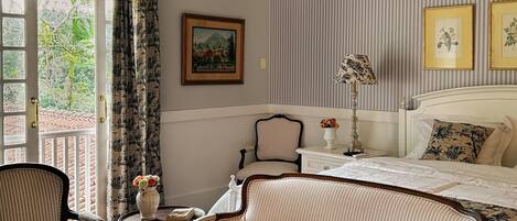 A room with a bed, a sofa, a chair, a table, and a chandelier. There is a window with curtains, a painting on the wall, and an air conditioning unit.