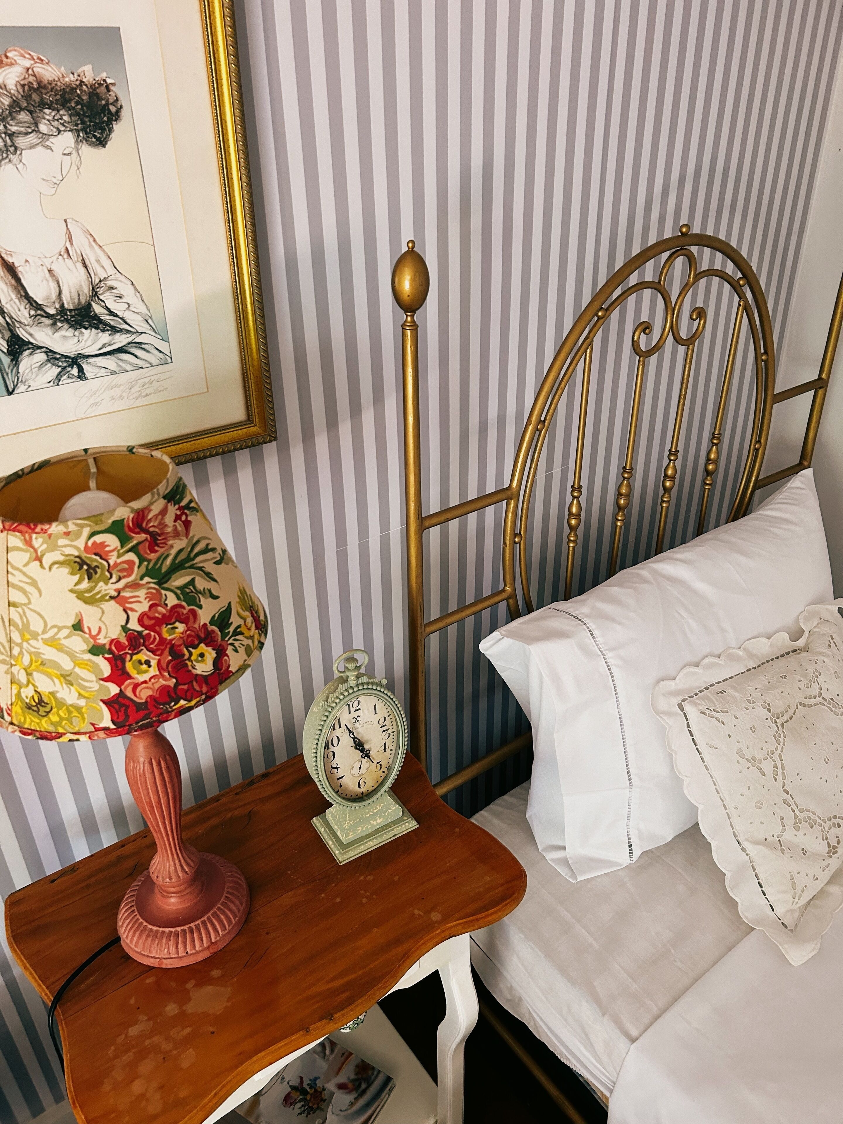 Una cama con ropa blanca, cabecera de latón, mesita de noche de madera con lámpara floral y un reloj antiguo.