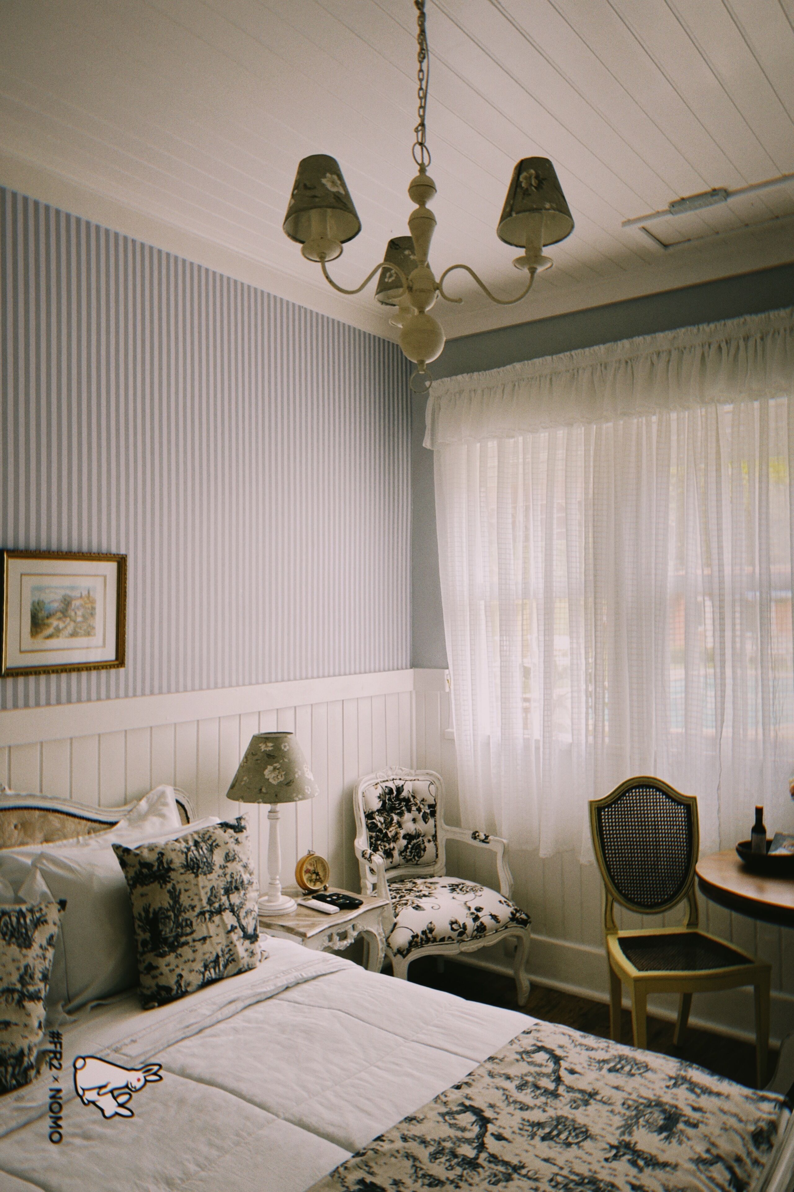 A room with a bed, two chairs, a table, a lamp, and a chandelier.