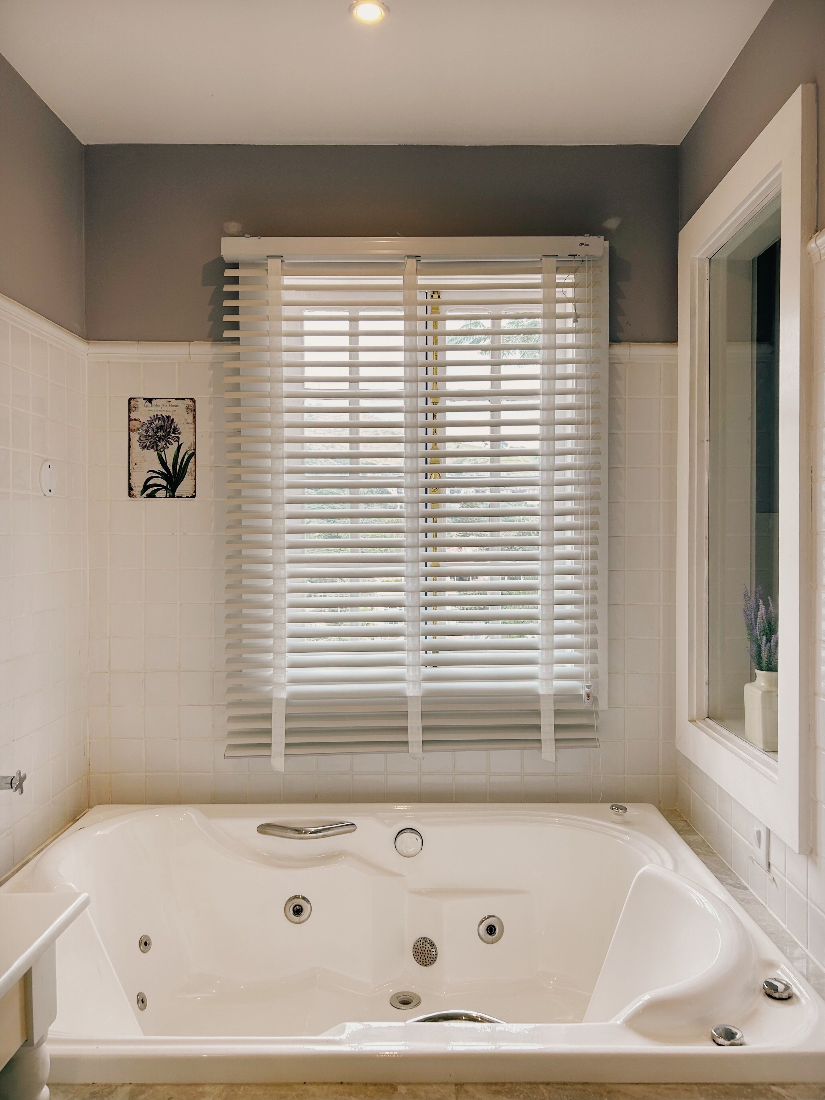 A bathroom with a bathtub, a window with blinds, and a wall-mounted air conditioning unit.