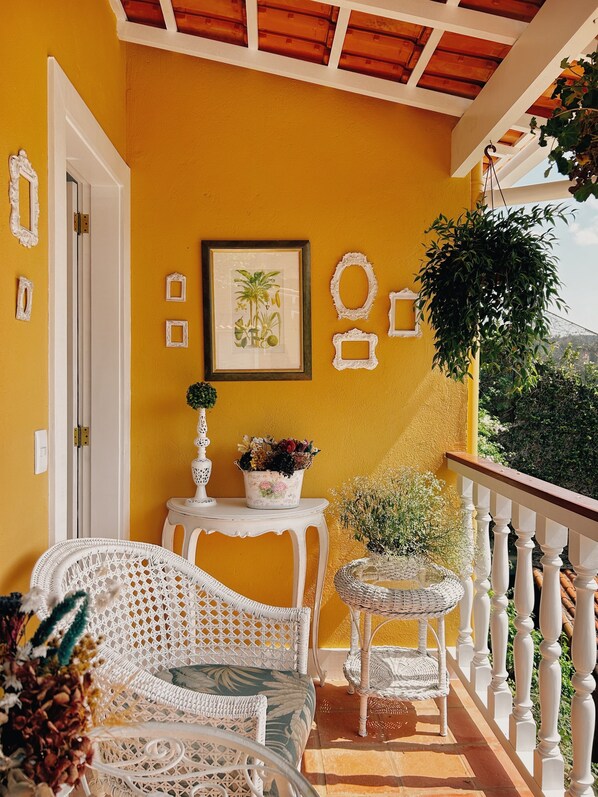 A balcony with white wicker furniture, a framed botanical print, and potted plants. - Hotel La Belle Provence (Petrópolis)
