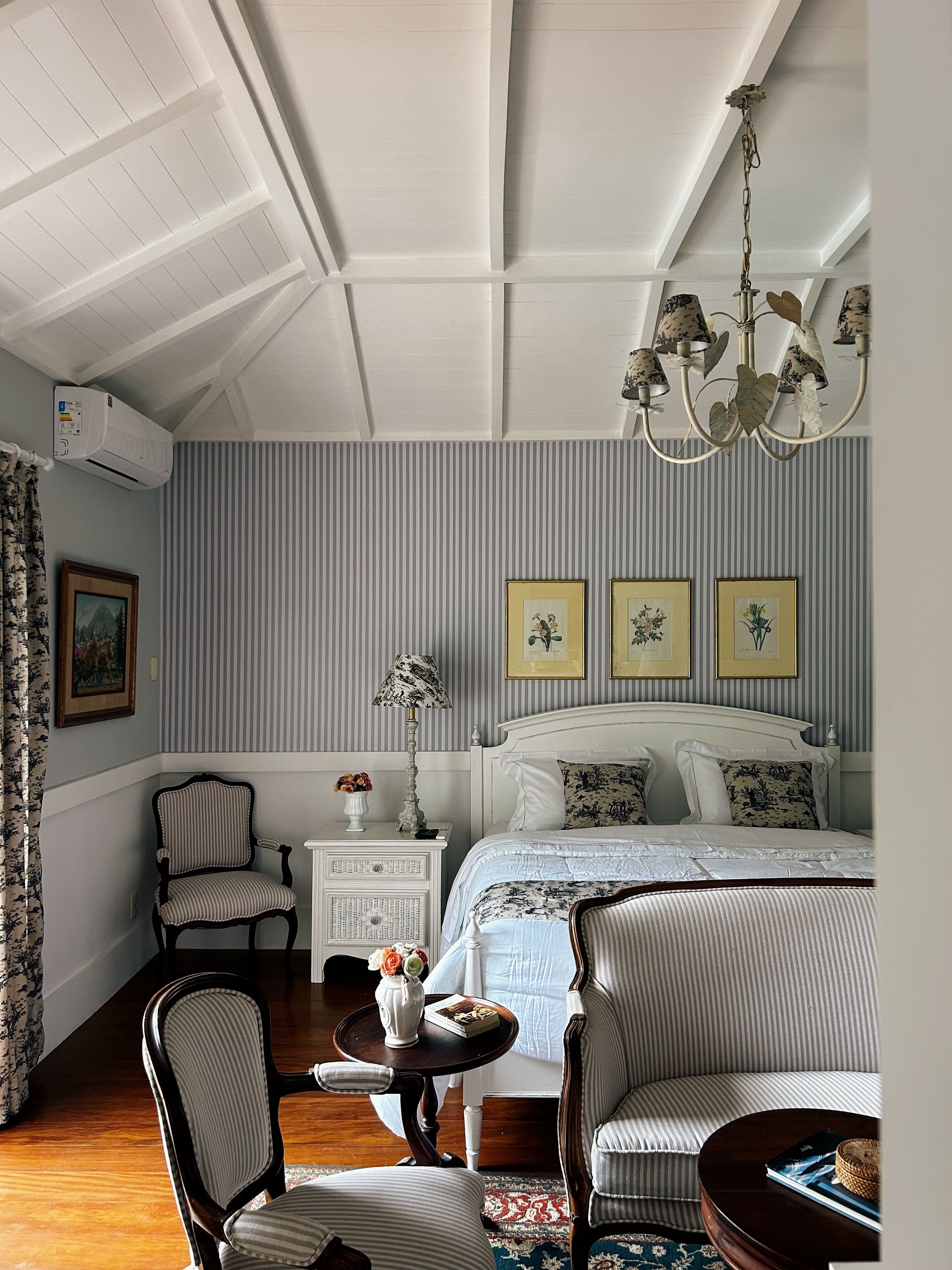 A bedroom with a bed, a chair, a nightstand, a lamp, and a chandelier.