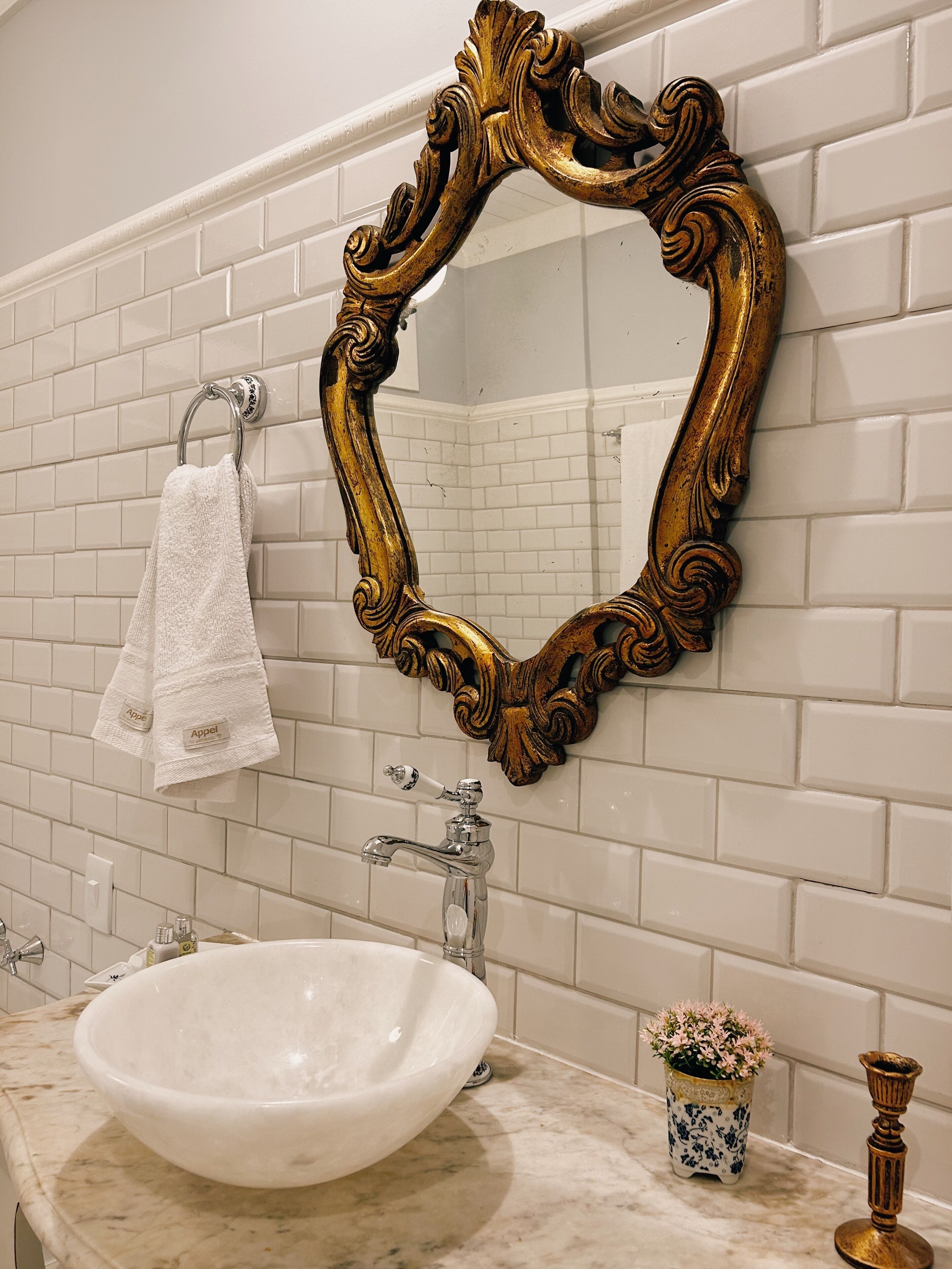 A bathroom with a white tiled wall, a gold ornate mirror, a white sink, and a small potted plant.