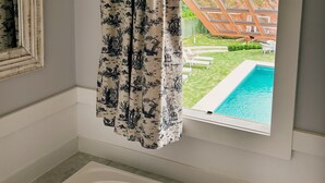 Un baño con bañera, ventana con vistas a una piscina y jardín, y cortina con diseño floral.