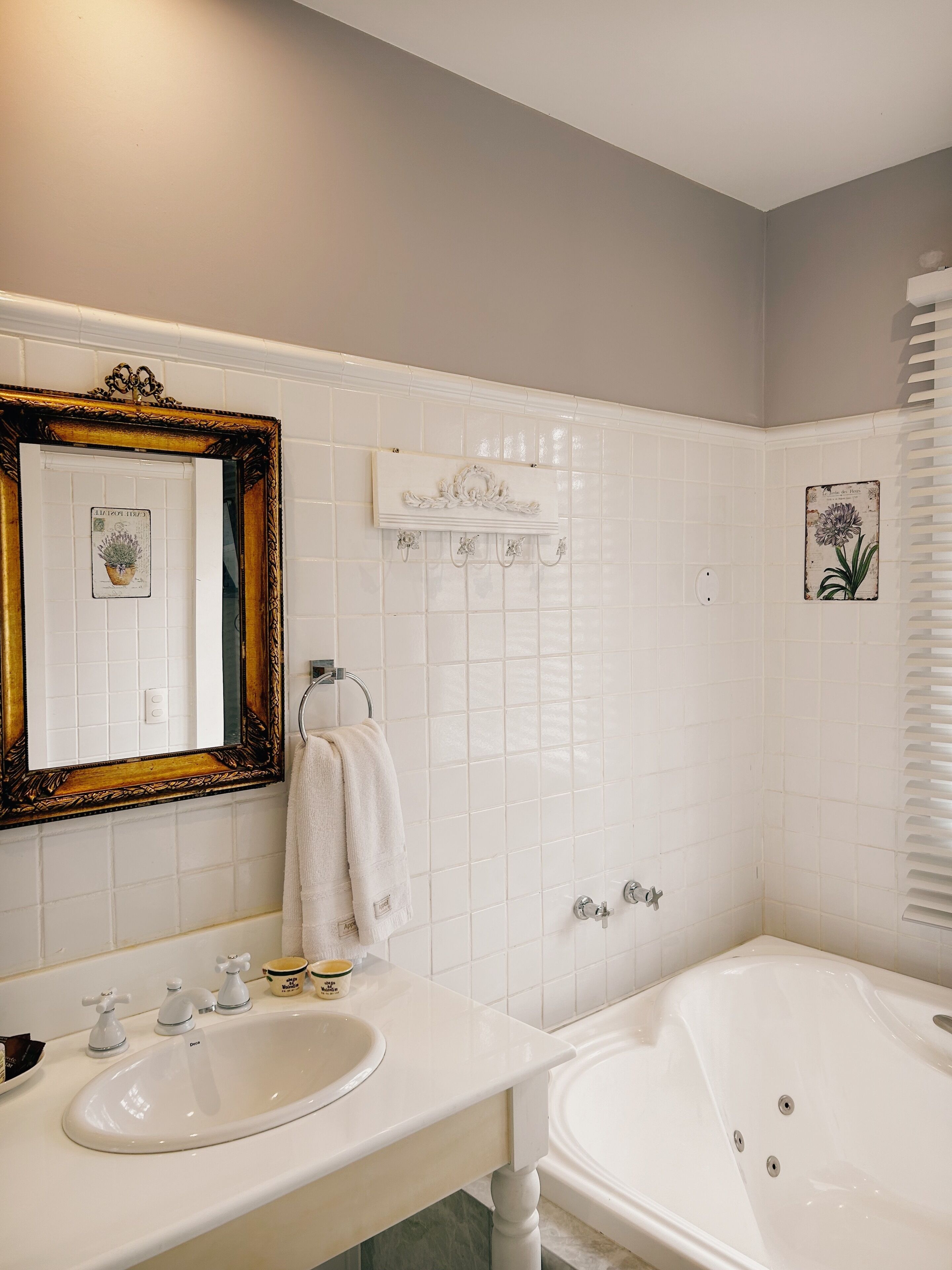 Um banheiro com pia, bancada e banheira brancos. Há um espelho com moldura decorada, porta-toalhas e um azulejo decorativo com motivos florais.