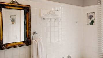 Um banheiro com pia, bancada e banheira brancos. Há um espelho com moldura decorada, porta-toalhas e um azulejo decorativo com motivos florais.