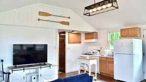 Cocina compacta con muebles blancos, estufa y refrigerador. Un televisor está montado en la pared, y hay un sofá azul en la sala.