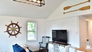 Una sala de estar con televisor, un sillón mecedor, un sofá azul, una rueda de barco de madera y un candelabro en el techo.
